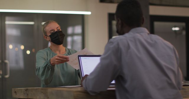 Two coworkers in a modern office; one hands documents to the other. They are practicing COVID-19 safety measures by wearing masks. One uses a laptop while engaged in discussion. Useful for business, teamwork, collaboration, and office environment themes.