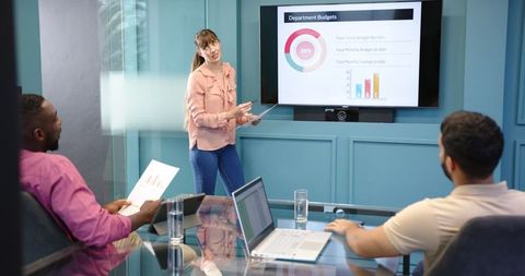 image features a woman presenting a department budget report on screen to colleagues in a modern office meeting room. this setting showcases teamwork and corporate collaboration, ideal for illustrating concepts such as business analysis, strategy sessions, and professional presentations in marketing materials or educational resources on business leadership.