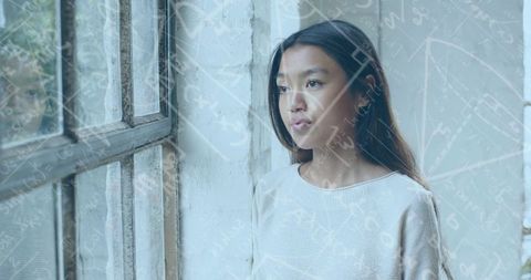 Contemplative Chinese woman by window wearing cream sweater with math formula overlay