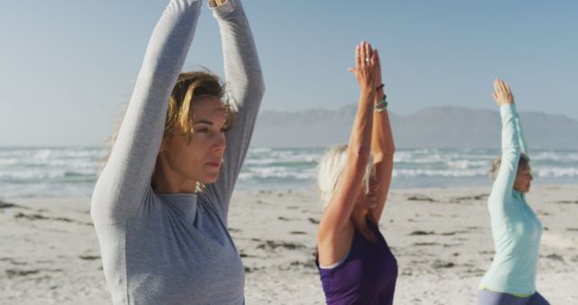 Group of women standing on a beach with arms raised overhead, practicing yoga by the ocean. This image is perfect for promoting outdoor fitness classes, wellness retreats, healthy living, and exercise routines in a natural setting.