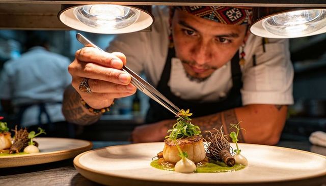 Chef plating seared scallop with microgreens and edible flowers using tweezers at pass