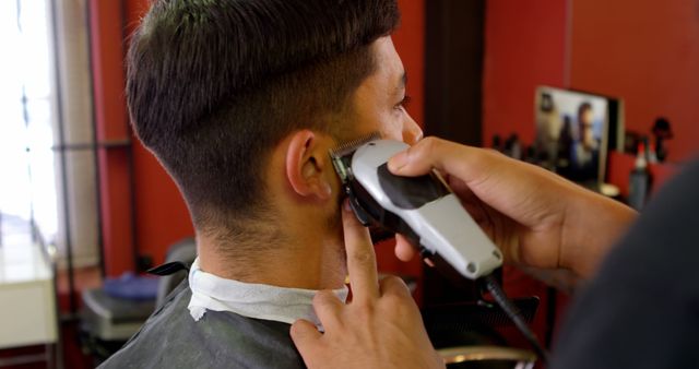 Man sitting in barbershop chair receiving a haircut with electric clippers. Ideal for use in grooming, styling, men's fashion, barbering services, and professional care contexts.