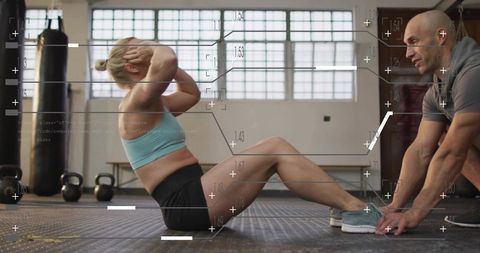 Woman performing seated crunches while trainer holding feet in urban gym with kettlebells
