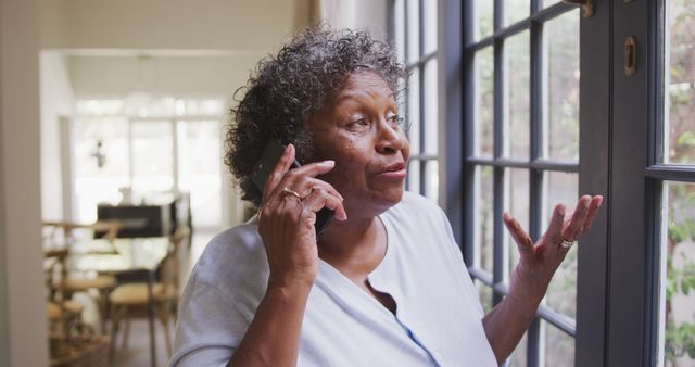 Senior african american woman looking through window and talking on smartphone. Senior lifestyle, free time, communication and domestic life.