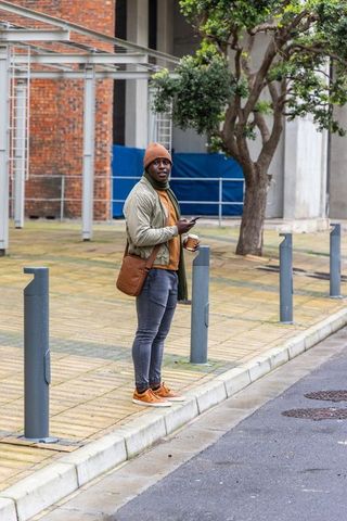 African american man standing on curb holding smartphone and coffee in urban plaza