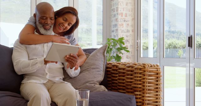 A cheerful African American couple is spending quality time together at home. The elderly man holds a tablet while the woman affectionately hugs him from behind. They are sitting on a comfortable sofa near large windows, enhancing the bright and airy atmosphere. Ideal for use in advertisements, websites, or articles focusing on personal technology, senior living, and family bonding.