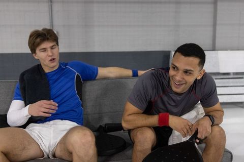 Two male athletes sitting and talking on bench holding pickleball paddles during court break