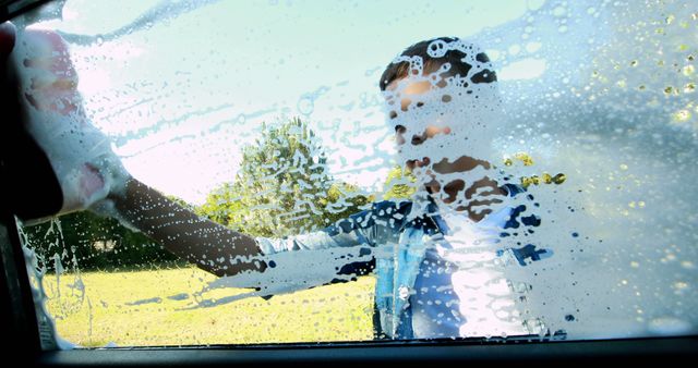 A young Caucasian boy is engaging in a car wash, playfully pressing his hand against the soapy window, with copy space. His cheerful interaction creates a sense of fun and innocence during an everyday task.