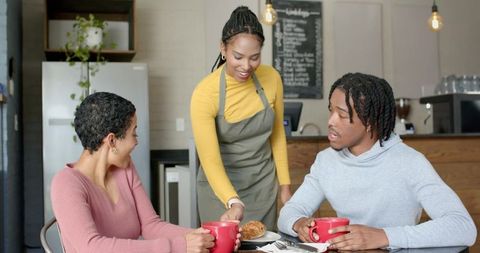 Barista serving pastry and coffee to smiling patrons holding red mugs in cozy urban cafe