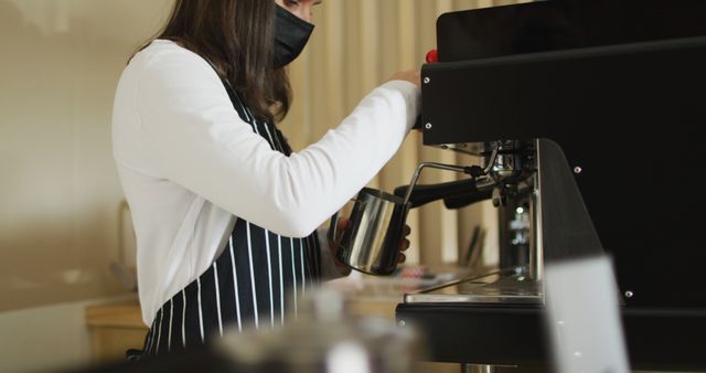 Female barista wearing striped apron and mask making coffee with espresso machine. Ideal for use in content related to coffee shops, barista training, professional coffee making, small business, and public health measures.