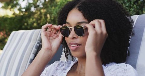 African american woman adjusting sunglasses on striped lounge chair in sunlit garden