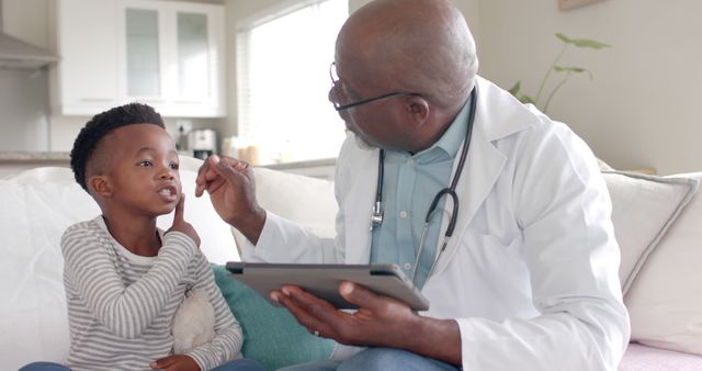 Older doctor in white coat using tongue depressor and tablet to check young boy's throat during home visit. Boy holding stuffed animal and opening mouth. Ideal for illustrating pediatric healthcare, medical consultations at home, and doctor-patient interactions in personal settings.
