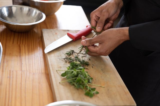 Chef is preparing fresh herbs on a wooden chopping board in a professional restaurant kitchen. This image is ideal for use in culinary blogs, cooking tutorials, restaurant websites, and food industry advertisements. It highlights the precision and skill involved in professional cooking.