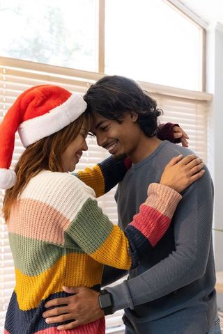 Couple sharing cozy holiday embrace by window wearing santa hat and knit sweaters