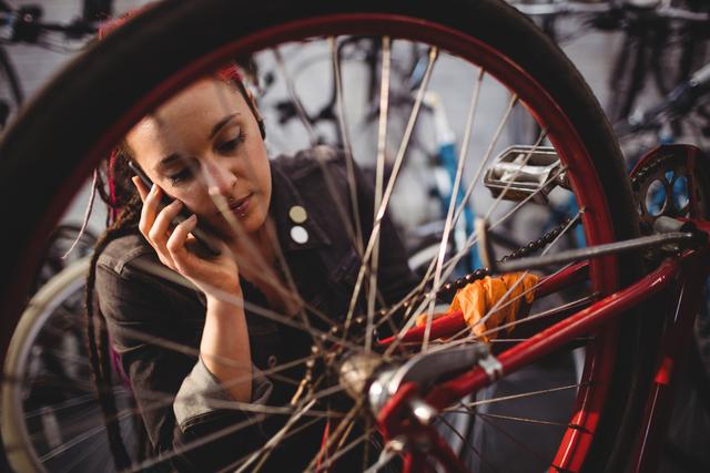 Mechanic multitasking by talking on phone while repairing bicycle in workshop. Useful for illustrating concepts of multitasking, professional repair services, and mechanical work. Ideal for articles, advertisements, and websites related to bike repair, maintenance services, and professional workshops.