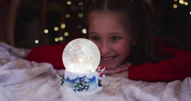 Child closely watching and admiring a lit Christmas snow globe featuring a Santa Claus figure against a backdrop of warm holiday lights. Great for holiday season promotions, greeting cards, advertisements, and Christmas-themed blog content.
