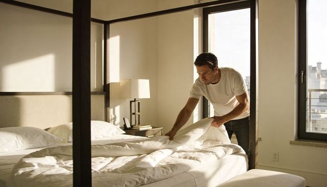 Man making bed in sunlit minimalist bedroom straightening white duvet near city windows