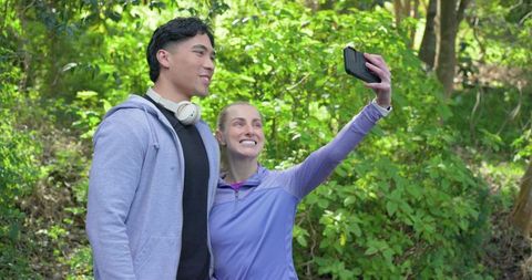 Smiling diverse couple taking selfie on sunlit forest trail during outdoor exercise