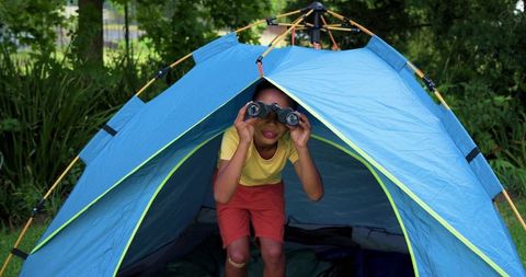 Curious child exploring backyard campsite with binoculars inside bright blue tent