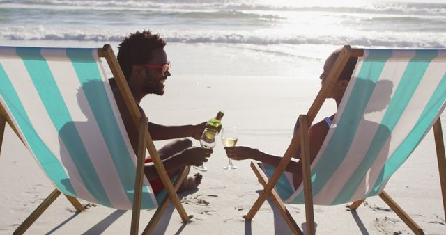 African american couple drinking wine together sitting on deck chairs at the beach. travel romantic vacation lifestyle concept