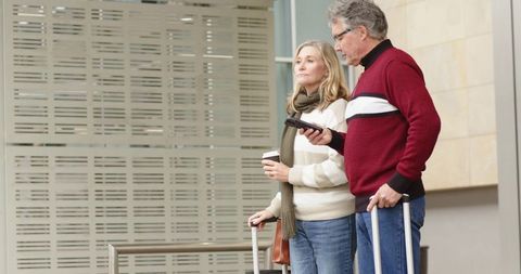 Mature couple waiting at airport concourse with luggage and coffee using smartphone