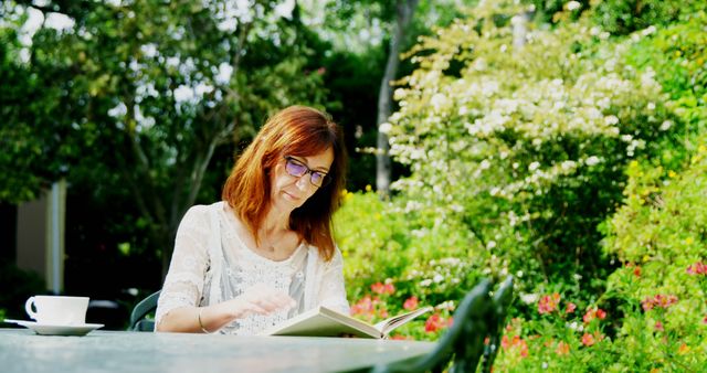 Woman sitting outdoors in a lush garden setting, engaged in reading a book. She is wearing glasses and appears to be enjoying the tranquility and nature around her. This is perfect for themes of relaxation, outdoor leisure activities, nature, and personal time.