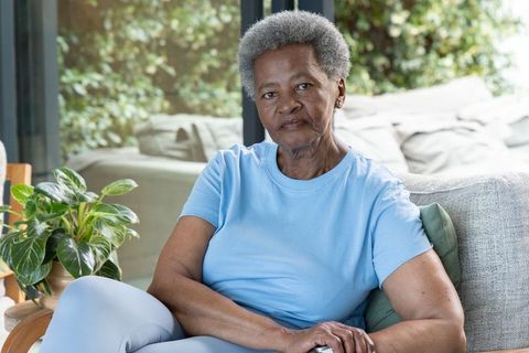Senior african woman sitting on sofa with hands clasped, calm living room portrait with greenery
