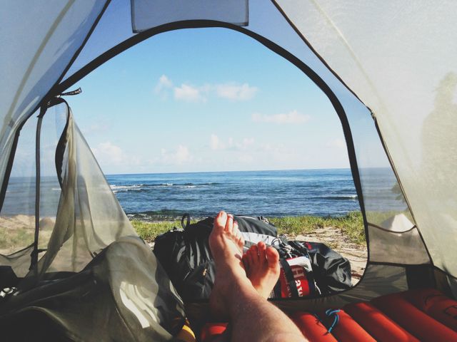 Person relaxing in tent, looking out at ocean. Ideal for travel blogs, outdoor adventure ads, beach resort promotions, vacation planning guides.