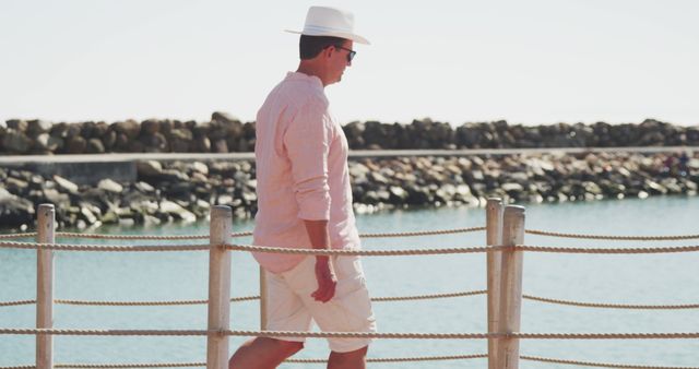 A man in light summer clothing, including a hat and sunglasses, walks near a beautiful coastal waterfront with a rocky shore in the background. This image can be used for travel blogs, vacation brochures, fashion websites, lifestyle articles, or outdoors and leisure advertisements.