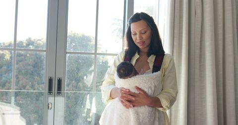 Asian american mother cradling swaddled infant by window with natural light