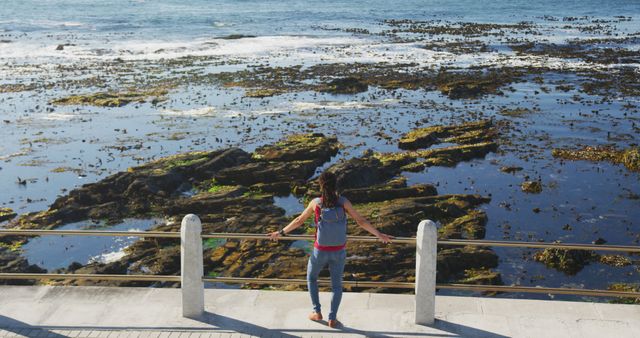 Woman in blue jeans with backpack enjoying a scenic view of the ocean from a coastal promenade. Ideal for travel blogs, lifestyle articles, and promotional content on tourism, nature, and outdoor activities.