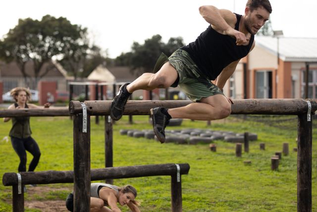 Front view of a Caucasian man wearing sports clothes in mid air, vaulting over a tall wooden hurdle at an outdoor gym during a bootcamp training session, with two women climbing over and under hurdles in the background