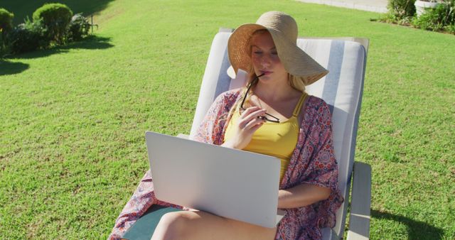 Woman sitting on lounge chair outdoors, wearing sun hat and patterned cover-up, working on laptop. Great for illustrating remote work, summer relaxation, outdoor leisure activities, freelance lifestyle, balancing work and relaxation.