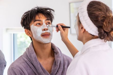 Diverse couple applying facial masks at home wearing bathrobes, relaxing skincare routine