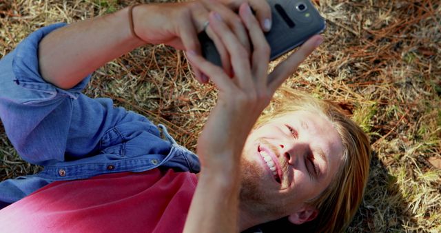 Young man lying on ground, smiling and using smartphone in natural outdoor setting. Perfect for depicting outdoor lifestyle, technology usage, relaxation, and leisure activities. Ideal for social media campaigns, advertisements showcasing digital device benefits, or content promoting outdoor experiences and youth connectivity.