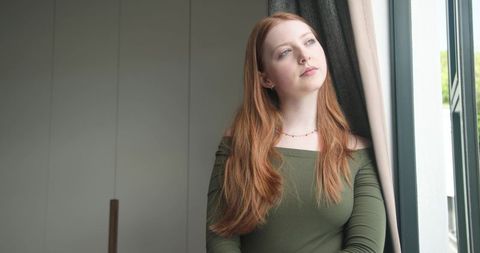 Red-haired woman gazing out window in olive off-shoulder top, sunlit contemplative portrait