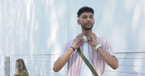 Young man adjusting headphones on sunlit urban sidewalk with crossbody bag