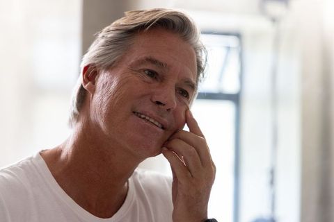 Thoughtful mature man resting hand on cheek near bright window, relaxed senior portrait