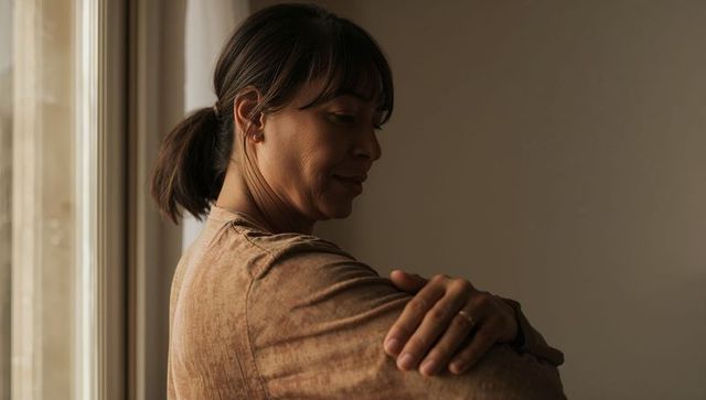Contemplative woman standing by sunlit window wearing brown top and ring, minimal interior