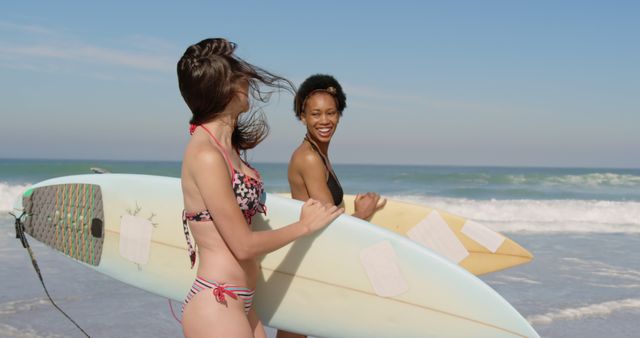 Two women wearing swimsuits are carrying surfboards while walking along a sunny beach. The ocean waves are crashing in the background. This image can be used for topics related to summer activities, beach vacations, surfing enthusiasm, leisure time, and friendships.