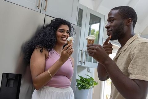 Diverse couple sharing vanilla ice cream cones, laughing, leaning on fridge in modern kitchen