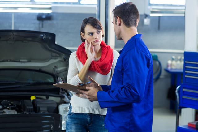 Mechanic in blue uniform discussing car repair checklist with female customer in auto repair garage. Ideal for illustrating automotive services, customer service interactions, vehicle maintenance, and professional consultations in repair shops.