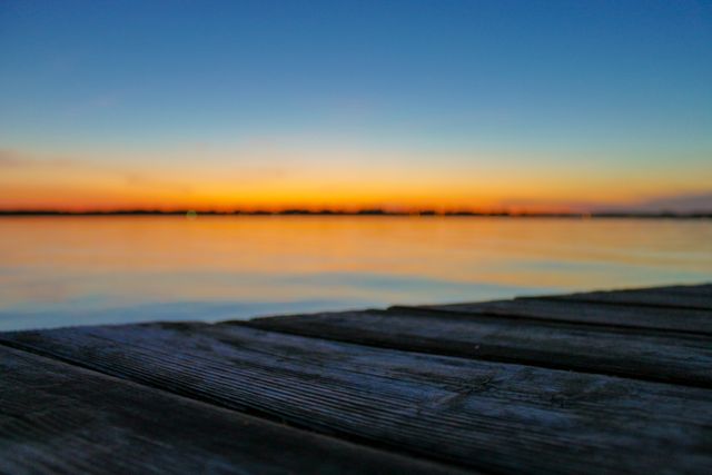 Wooden pier stretching out towards calm lake during sunset, with sky painted in colorful hues and horizon softly blending with the water. Ideal for use in travel brochures, relaxation-themed designs, desktop wallpapers, and nature blogs emphasizing tranquility and natural beauty.