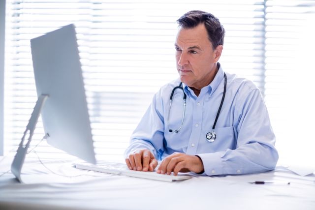 Male doctor in clinic working on computer, wearing stethoscope around neck. Ideal for use in healthcare, medical technology, telemedicine, and professional work environment contexts.