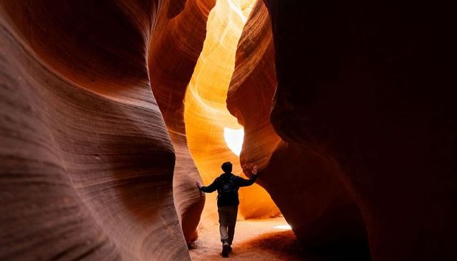 Solo hiker navigating sunlit narrow slot canyon with flowing sandstone layers