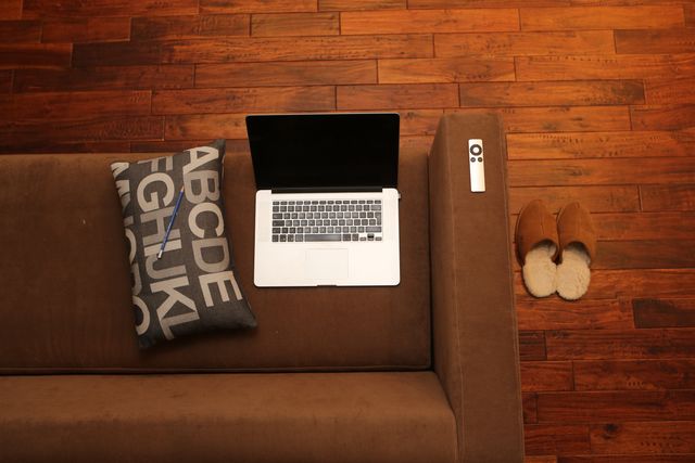 This image depicts a home office environment with a laptop on a sofa, a remote control, and comfortable slippers on a wooden floor. Ideal for illustrating modern work-from-home setups, remote working, and cozy interior concepts for blogs, advertisements, or articles emphasizing work-life balance and home comfort.