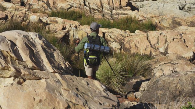 An African American man is navigating a rocky landscape with trekking poles and a backpack, showcasing themes of outdoor adventure and fitness. This is ideal for use in content related to hiking adventures, encouraging an active lifestyle, or travel brochures focused on ecotourism and exploration.