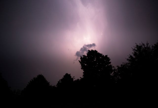 Dramatic scene with lightning illuminating the sky against the dark silhouettes of trees during a storm. Useful for themes related to nature's power, weather, and dramatic atmospheric conditions, or as a background for storytelling involving storms and intense weather events.