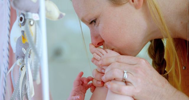 A close-up view of a mother with blond hair tenderly kissing her baby's feet in a nursery environment. Perfect for advertising parenting products, illustrating motherhood segments in magazines, or showcasing nurturing family moments.