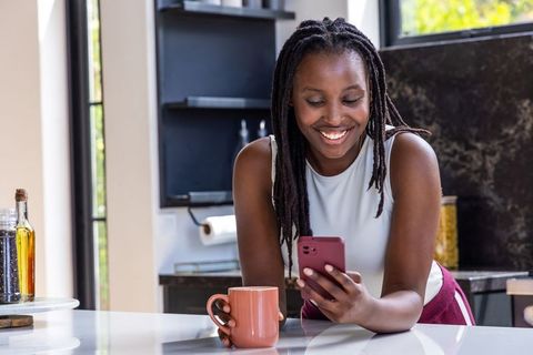 Smiling woman checking smartphone while leaning on modern kitchen island holding coffee mug